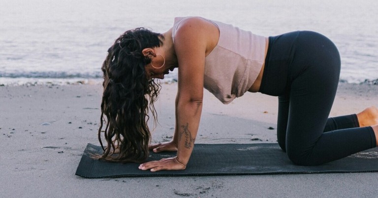 Frau in der Kuh Yogahaltung (Bitilasana) am Strand auf Yogamatte