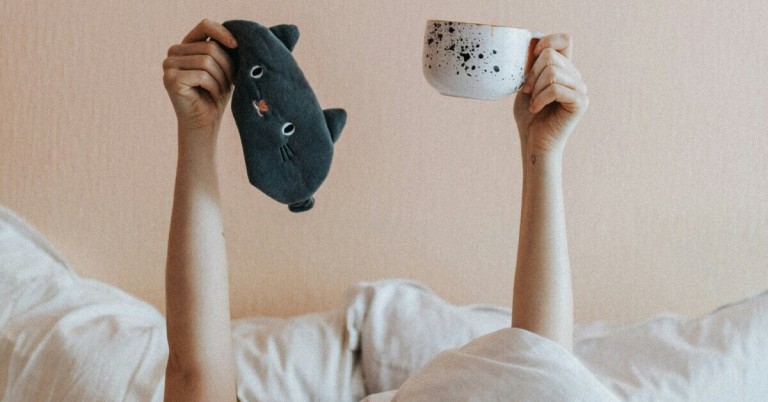 Person hält eine Tasse Kaffee und eine Schlafmaske im Bett – Kaffee vor dem Schlafen