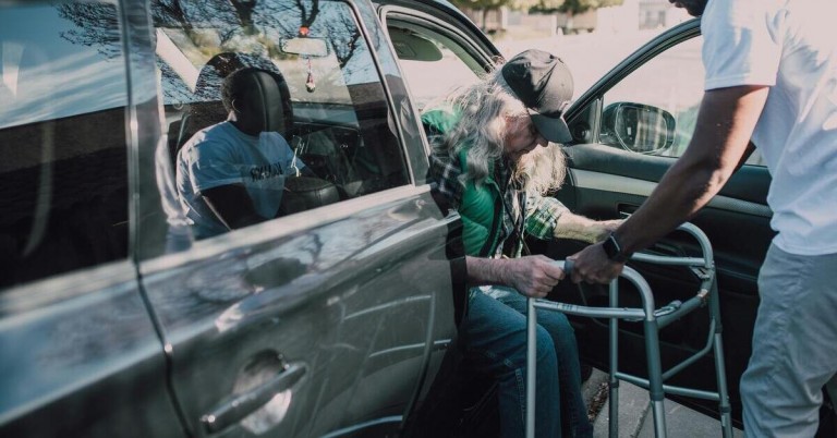 Älterer Mann steigt mit Hilfe eines Gehgestells und Unterstützung aus einem Auto aus.