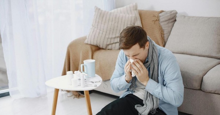Mann mit Erkältung sitzt auf dem Sofa, benutzt Taschentuch und hat Medikamente auf dem Tisch